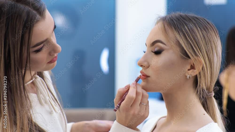 Young brunette woman make-up artist paints a blonde girl in a beauty salon. Paints lips. Young brunette woman make-up artist paints a blonde girl in a beauty salon. Paints lips.