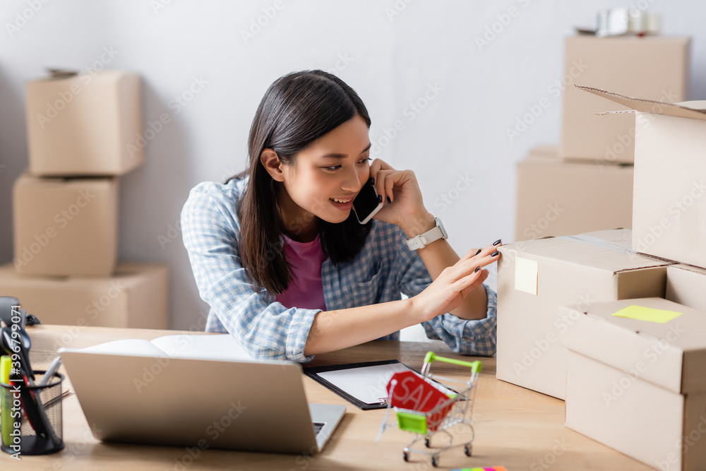Smiling asian volunteer talking on smartphone while touching carton box on desk in charity center on blurred background Smiling asian volunteer talking on smartphone while touching carton box on desk in charity center on blurred background