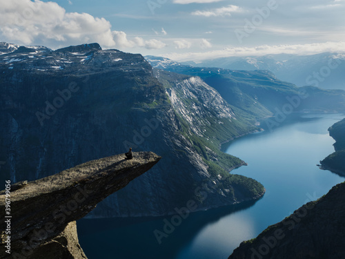 Hiker sitting on the Trolltunga in Norway with a view over the deep blue fjord below and a montain range in the background