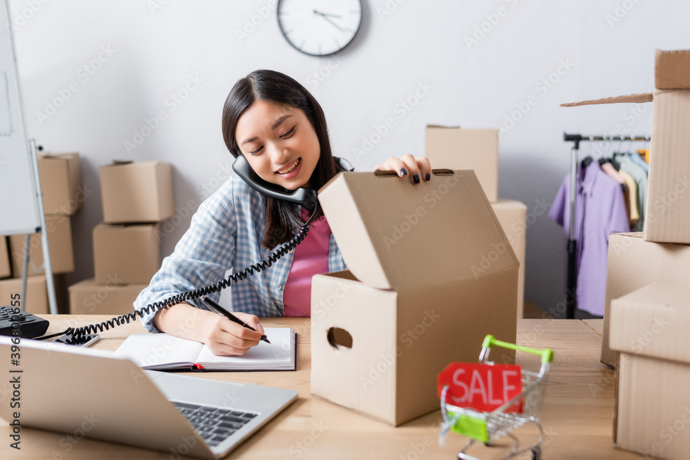 Smiling asian volunteer talking on telephone and writing in notebook while opening carton box at desk in charity center Smiling asian volunteer talking on telephone and writing in notebook while opening carton box at desk in charity center