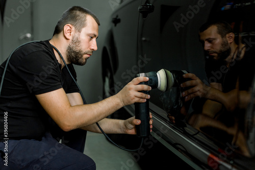 Car detailing and polishing concept. Professional Caucasian male car service worker, wearing black uniform, holding in hands orbital polisher, and polishing body of black luxury car