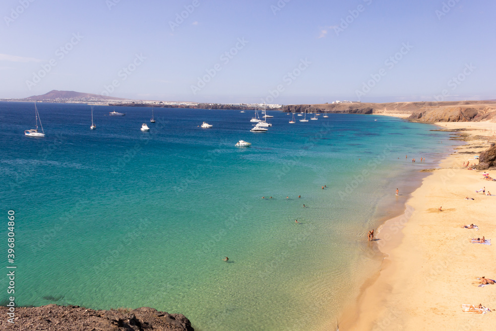 Few people on Papagayo beach with sailboats over turquoise water sea in Lanzarote. Tourist bay on volcanic coast in Canary Islands on sunny day. Summer holidays, travel destination concepts