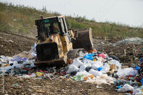 Yellow tractor on dumping site. Plastic and paper garbage. Waste sorting on landfill of big city. Caring for environment.