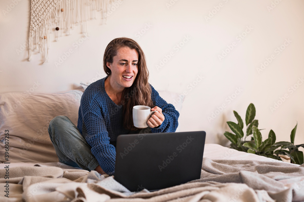 Woman sat in bed drinking mug of coffee and using laptop Woman sat in bed drinking mug of coffee and using laptop