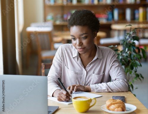 Millennial black woman studying or working online, taking notes in copybook at urban cafe