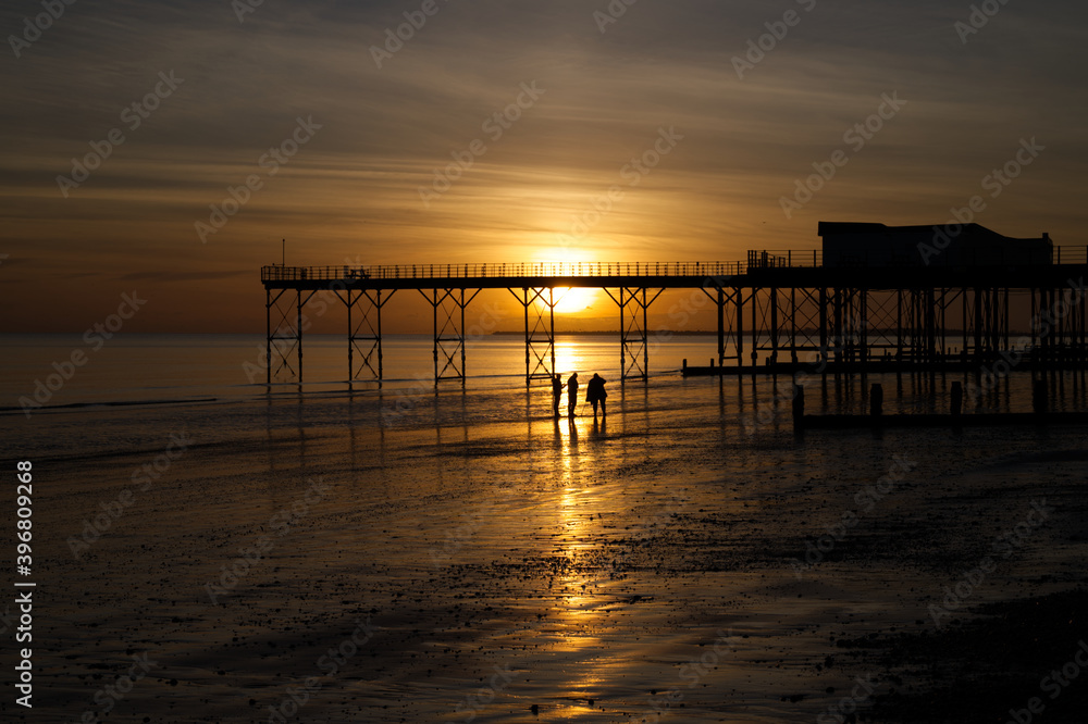 A stunning sunset behind the Pier at Bognor Regis with beautiful reflections of the sinking sun in the sea and sand with locals admiring the spectacle. 