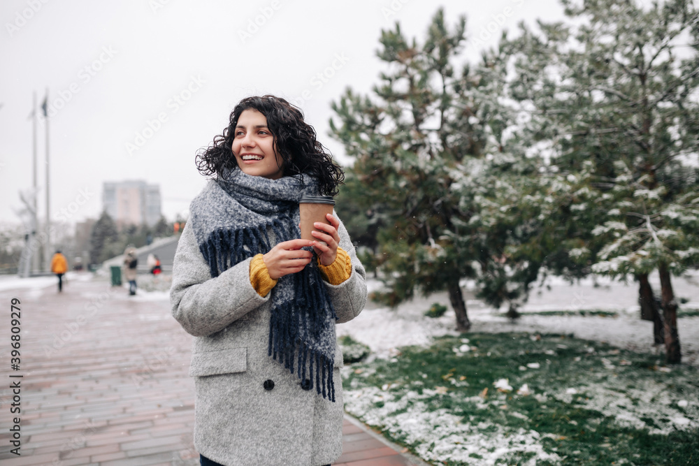 Young woman wearing frey fashion coat and blue scarf stands with a coffee to go at a winter snowy park. Female enjoys hot drink during frosty cold day outdoors. Joy and holidays concept.
