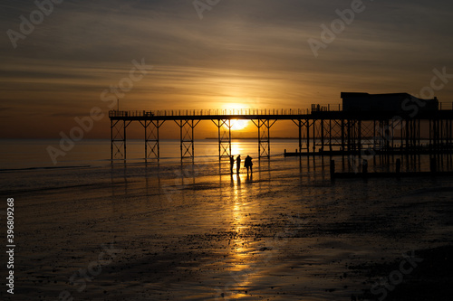 A stunning sunset behind the Pier at Bognor Regis with beautiful reflections of the sinking sun in the sea and sand with locals admiring the spectacle. 