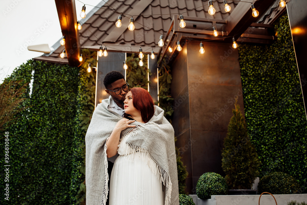 Portrait of beautiful couple wrapped in blanket stand at the balcony, handsome African American man hug gorgeous white woman, enjoy winter holidays, Christmas morning concept
