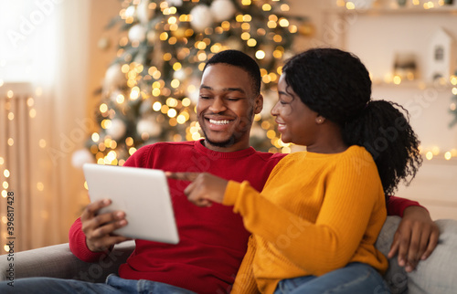 Happy millennial lady points finger at tablet and shows to guy something interesting in interior with tree and garlands