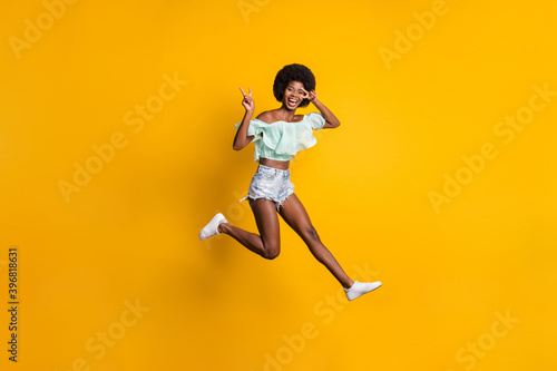 Full length body size photo of jumping smiling black skinned girl showing v-sign fingers isolated on vivid yellow color background