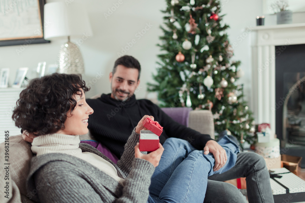 Wife opening Christmas gift from husband on living room sofa Wife opening Christmas gift from husband on living room sofa