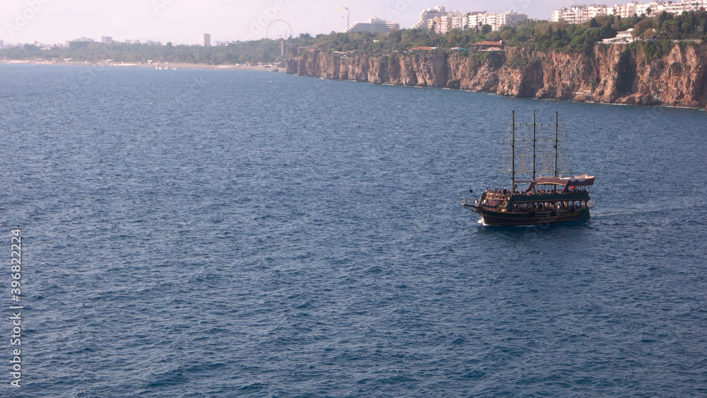Obraz premium Beautiful yacht with tourists floating on the sea. Blue wavy sea with resort town in the background. Summer vacation concept. Travel to Turkey.