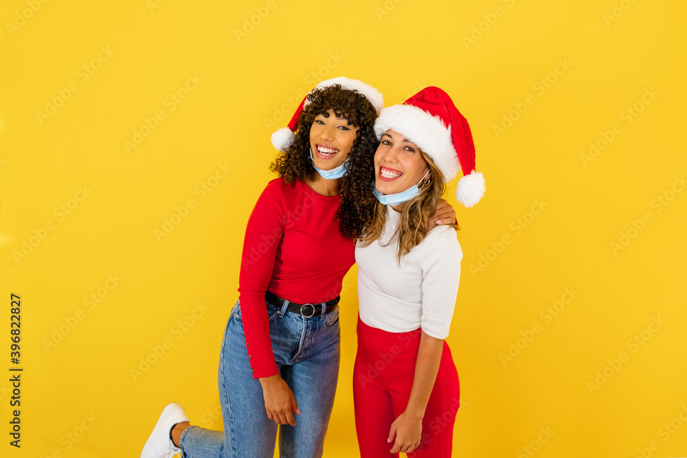 Beautiful latina curly brunette embracing her best female Caucasian friend smiling confident for future - Two mixed race women on yellow background in Santa hat wearing Coronavirus protection mask Beautiful latina curly brunette embracing her best female Caucasian friend smiling confident for future - Two mixed race women on yellow background in Santa hat wearing Coronavirus protection mask