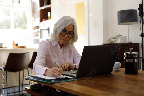Senior caucasian woman with laptop talking notes while working from home