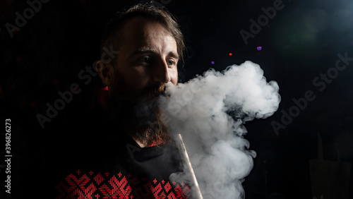 Close-up photo of man smoking traditional hookah pipe. Man exhaling smoke in hookah cafe on dark background. Hookah or water pipe concept.