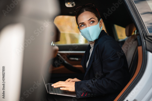 Focused businesswoman wearing face mask working with laptop in car