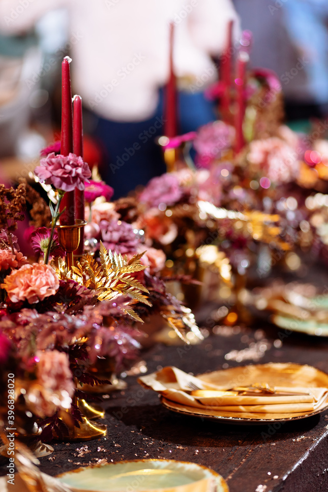 Christmas wedding dinner served table. Shiny leaves of gold paint, pink and red flowers and gold dishes. candles in a metal candlestick. Selected focus and blurred background