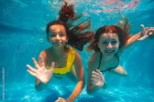 Two happy girls swim underwater and wave hand to the camera smiling in the pool