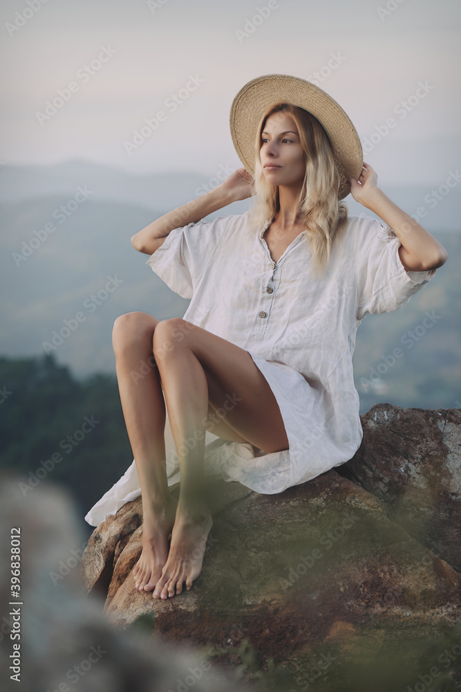 The blonde in a linen dress and straw hat sits on a viewpoint in the mountains. A woman in the Boho style, resting in the countryside, a simple village lifestyle