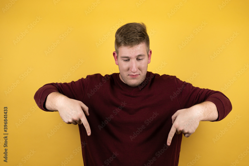 Young blonde man wearing a casual red sweater over yellow background surprised, looking down and pointing down with fingers and raised arms Young blonde man wearing a casual red sweater over yellow background surprised, looking down and pointing down with fingers and raised arms