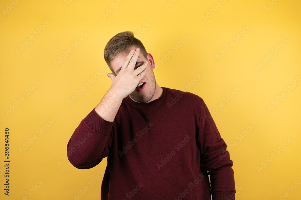 Young blonde man wearing a casual red sweater over yellow background peeking in shock covering face and eyes with hand, looking through fingers with embarrassed expression Young blonde man wearing a casual red sweater over yellow background peeking in shock covering face and eyes with hand, looking through fingers with embarrassed expression