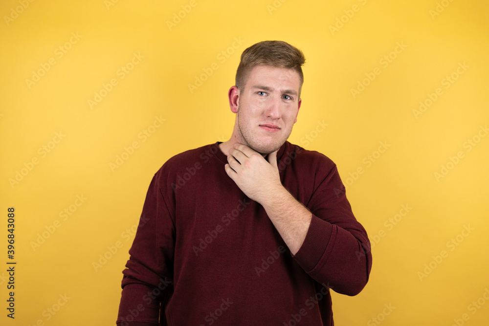 Young blonde man wearing a casual red sweater over yellow background touching painful neck, sore throat for flu, clod and infection Young blonde man wearing a casual red sweater over yellow background touching painful neck, sore throat for flu, clod and infection