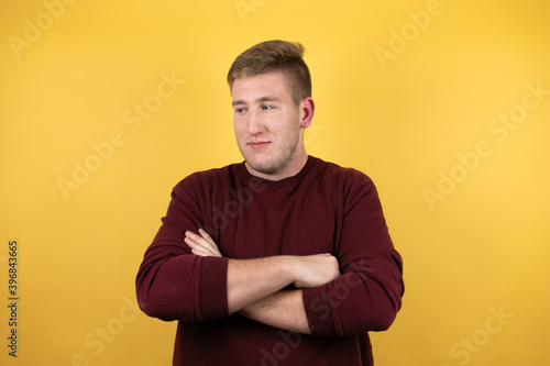 Young blonde man wearing a casual red sweater over yellow background thinking looking tired and bored with crossed arms