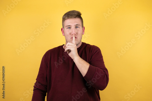 Young blonde man wearing a casual red sweater over yellow background asking to be quiet with finger on lips. Silence and secret concept.