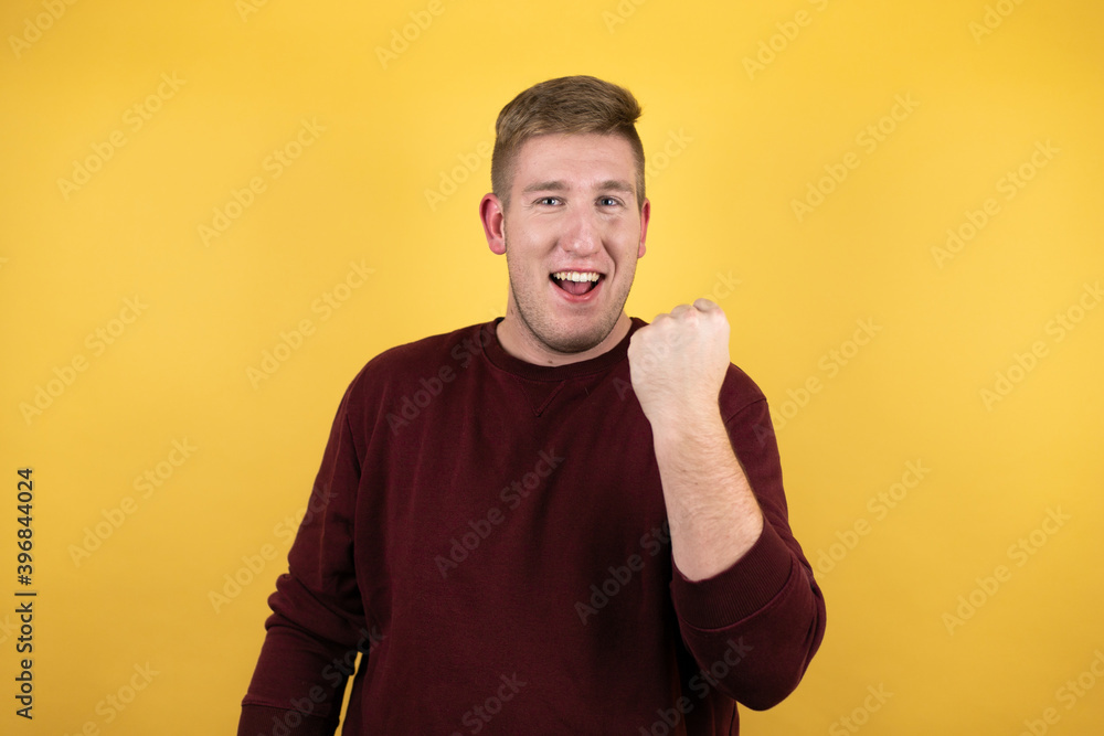 Young blonde man wearing a casual red sweater over yellow background angry and mad raising fist frustrated and furious while shouting with anger. Rage and aggressive concept. Young blonde man wearing a casual red sweater over yellow background angry and mad raising fist frustrated and furious while shouting with anger. Rage and aggressive concept.