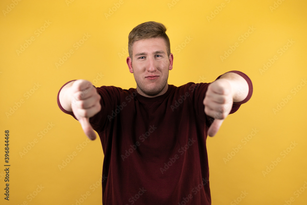 Young blonde man wearing a casual red sweater over yellow background doing negative sign showing dislike with thumb down Young blonde man wearing a casual red sweater over yellow background doing negative sign showing dislike with thumb down