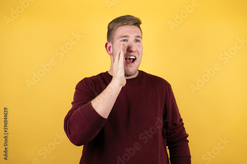 Young blonde man wearing a casual red sweater over yellow background shouting and screaming loud to side with hands on mouth