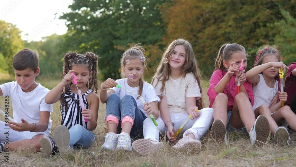 Cute happy kids in school age sitting together in park and blowing soap bubbles Cute happy kids in school age sitting together in park and blowing soap bubbles