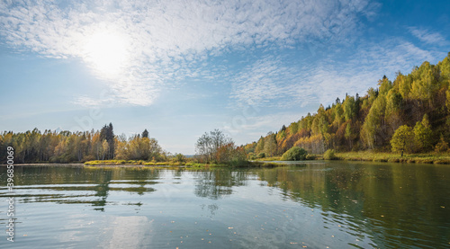 Autumn forest trees are reflected in the river water of the panoramic landscape. Blue sky with clouds.