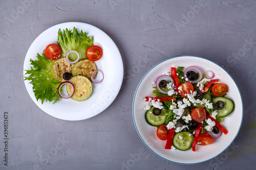 White plate with grilled zucchini with cherry tomatoes on lettuce leaves and greek salad on a concrete background.