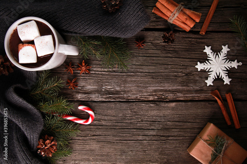 Cocoa with marshmallow, cinnamon, knitted scarf, bauble, fir tree branches, cones, craft gift box, candy cane on rustic wooden table. Winter and Christmas concept. Top view, copy space