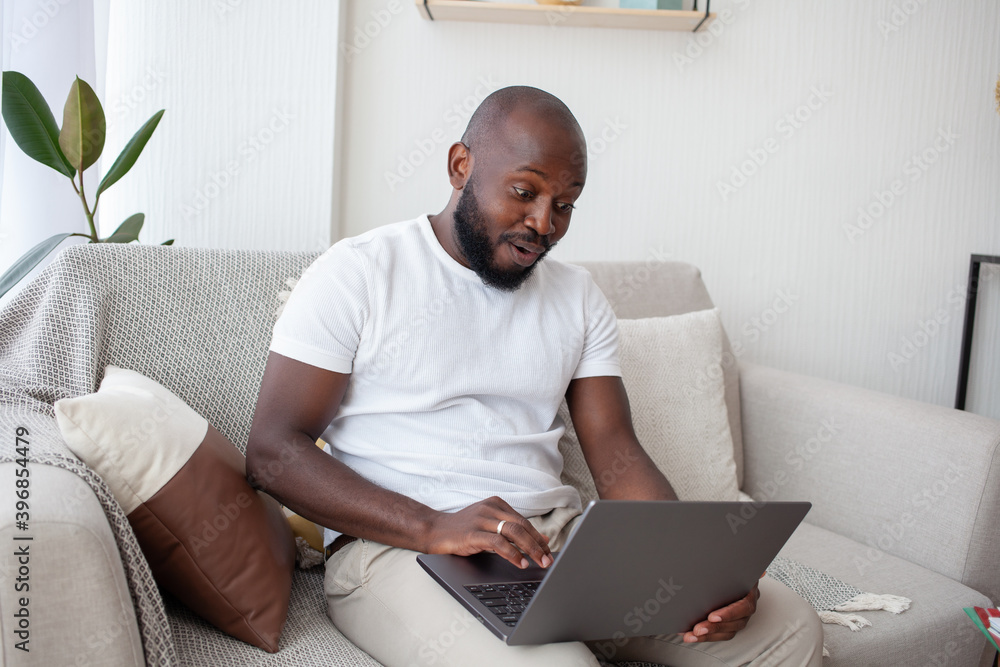 African-American man is pleasantly surprised, watching an exciting show on the Internet, relaxing sitting on gray sofa in living room. People, resting and TV concept. African-American man is pleasantly surprised, watching an exciting show on the Internet, relaxing sitting on gray sofa in living room. People, resting and TV concept.