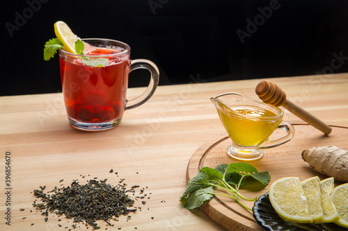 Berry tea in a cup next to lemon, honey, lemon balm, ginger and tea leaves on the table.