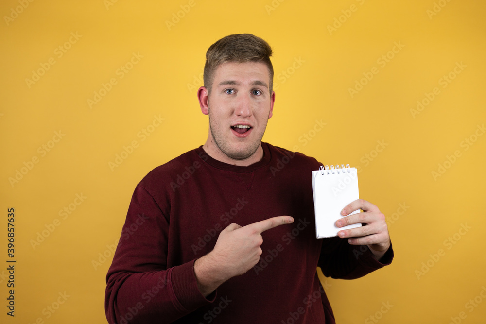 Young blonde man wearing a casual red sweater over yellow background pointing blank notebook in her hand Young blonde man wearing a casual red sweater over yellow background pointing blank notebook in her hand