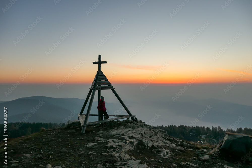 A man standing next to a small tower with a cross on the top on the peak of Gerlitzen, Austrian Alps and observing the daybreak. The skyline is exploding with orange. The valley is shrouded in fog