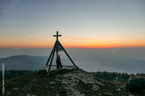 A man standing next to a small tower with a cross on the top on the peak of Gerlitzen, Austrian Alps and observing the daybreak. The skyline is exploding with orange. The valley is shrouded in fog