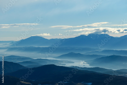 A panoramic view on Austrian Alps, captured on an early morning. The high peaks are hiding behind light clouds. The valley is shrouded in fog. There is a long lake at the bottom of the valley. Remedy © Chris