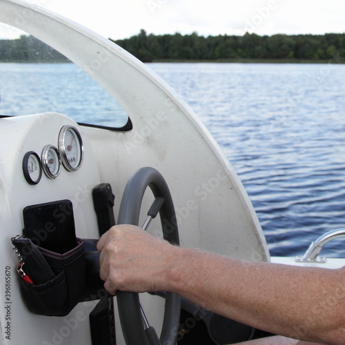 Man's hand on floating motor boat steering wheel, cabin watercraft interior dashboard with control devices at summer day on water and river shore background, outdoor travel on private boat recreation