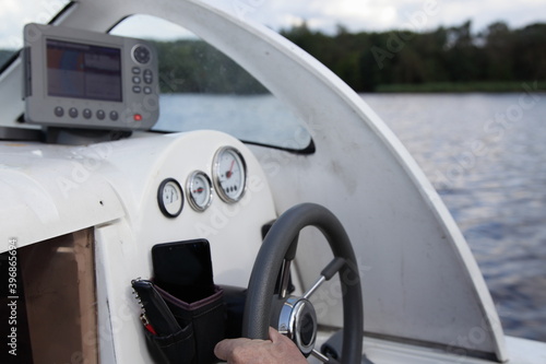 Man's hand on floating motor boat steering wheel, cabin watercraft interior dashboard with control appliances and chart plotter at summer day on water and river shore background, outdoor travel © Ilya