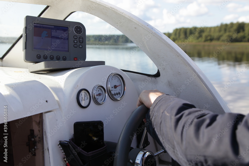 Man's hand in jacket on floating motor boat steering wheel, cabin watercraft interior dashboard with control devices at summer day on water and river shore background outdoor travel on boat recreation