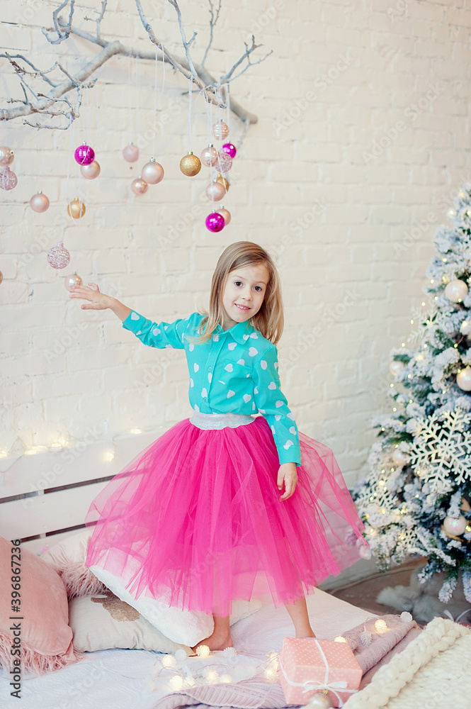 Little curious girl stands on a wooden chair to look closer at the Christmas toys hanging as decoration on the wall next to the Christmas tree in a white bright interior style. Xmas home decor concept Little curious girl stands on a wooden chair to look closer at the Christmas toys hanging as decoration on the wall next to the Christmas tree in a white bright interior style. Xmas home decor concept