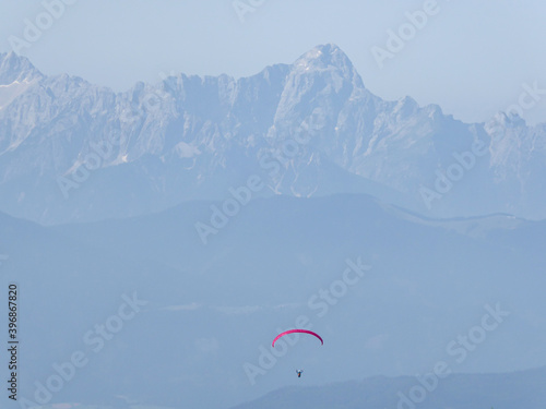 A paraglider floating in between Alpine peaks in Austria. The parachute is red. Free drifting. the the back there are steep and massive mountain chains. Dangerous sport. Adrenaline
