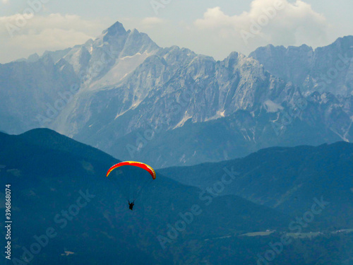 A paraglider floating in between Alpine peaks in Austria. The parachute is red. Free drifting. the the back there are steep and massive mountain chains. Dangerous sport. Adrenaline