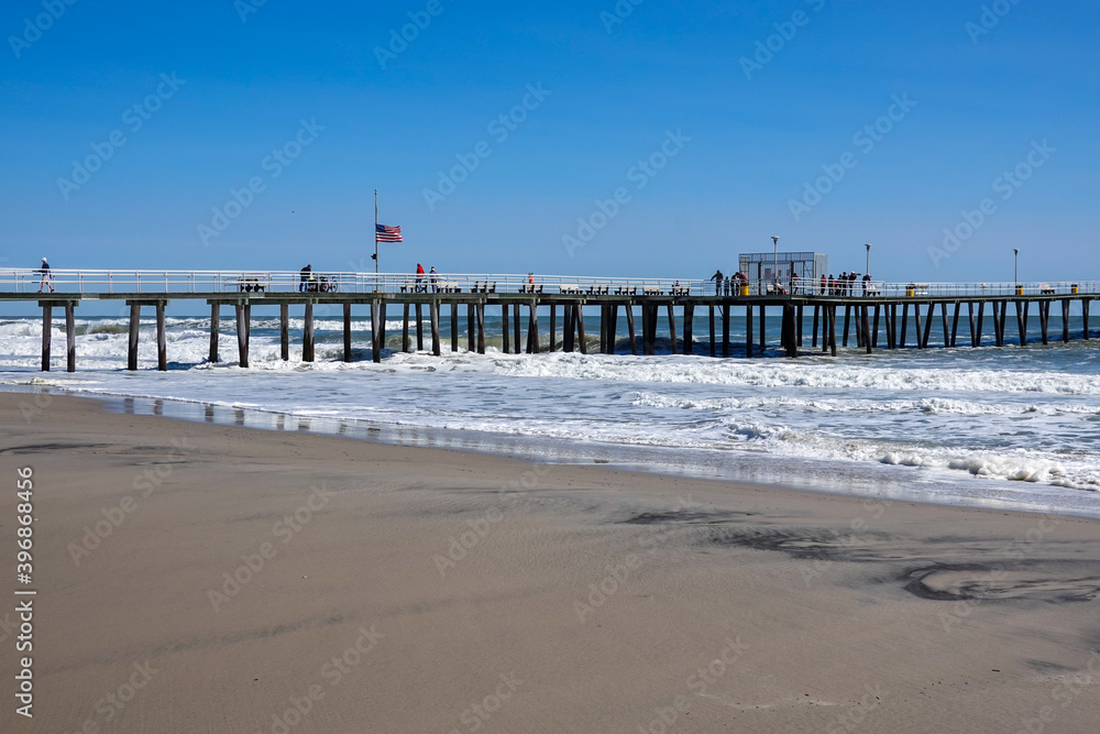 Rough ocean waves hit the wooden pylons of a long pier that has an American Flag and people walking on it