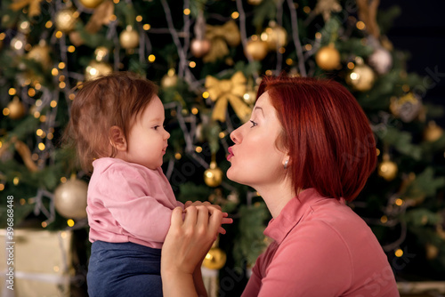 Mother holding hands and kissing infant baby daughter in front of Christmas (New Year) tree decorated with ligths and golden balls and bows. Gift boxes in background. Family winter holidays concept